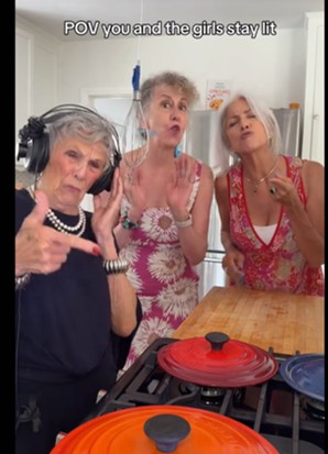 Three older women singing together in a kitchen in a Retirement House video, one wearing headphones and one holding a whisk like a microphone