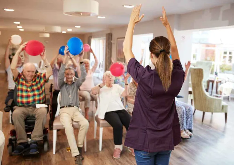 Activities coordinator leading a seated balloon activity for older residents, including a man in a power wheelchair, in a senior living community room