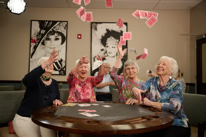 Four older women laughing as they toss playing cards into the air around a game table in a senior living lounge