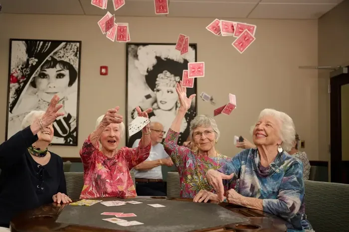 Four older women laughing as they toss playing cards into the air around a game table in a senior living lounge