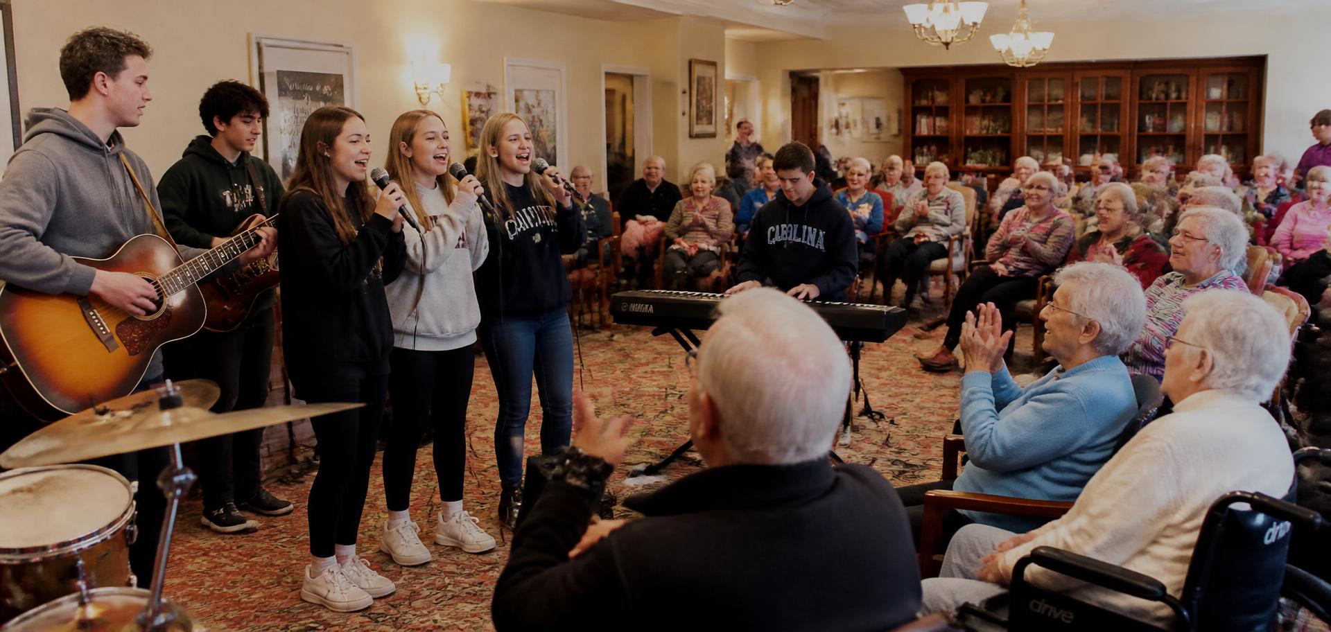 A group of student performers singing for a full room of senior residents